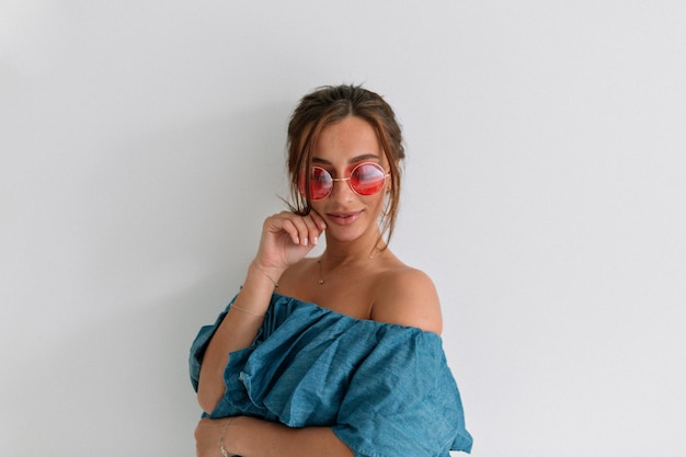 Close up photo of pretty trendy woman in round pink glasses touching her chin and looking down over isolated white wall