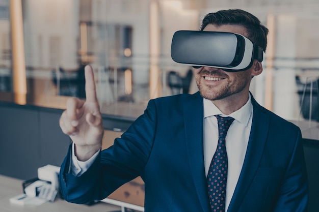 Close-up photo of smiling young bearded finacier in formal suit enjoying spending time in virtual reality with VR headset glasses, pointing with forefinger up in air. Cyberspace experience concept