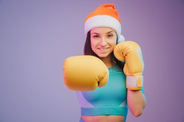 Close up photo of strong sportswoman in boxing gloves and Christmas hat, boxer posing. isolated on gradient studio background in neon light. Beautiful caucasian woman practicing.