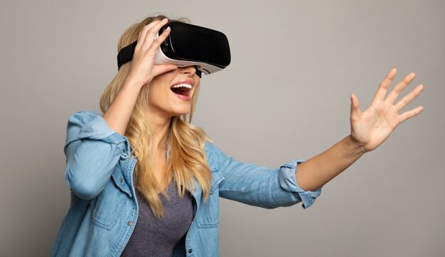 Close up photo of a stunned woman in a casual outfit, who is wearing VR glasses, holding them with her right hand and stretching her left hand towards the camera.