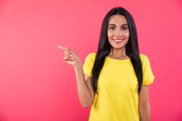 Close-up photo of a stunning happy girl, who is pointing to the left with her right index finger and posing in a yellow t-shirt on a pink background