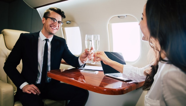 ビジネスクラスの飛行機のボードで飛行機に乗っている間、同僚とシャンパンのフルートでチャリンという音を立てている成功した男性のクローズアップ写真。