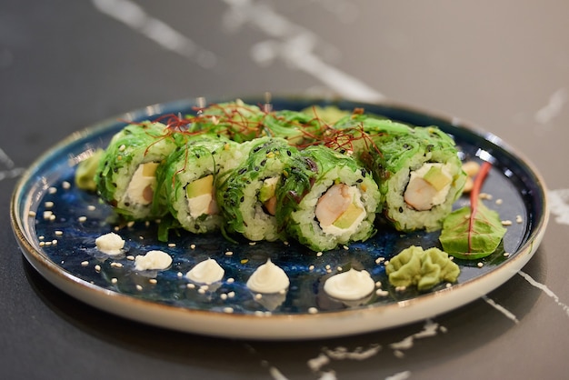 A close-up photo of sushi rolls with Chuka wakame seaweed, Philadelphia cheese, and shrimps on a blue ceramic plate with sauce, cashew, and wasabi on a black stone table.