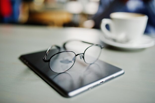 カフェテーブルのコーヒーのカップに対してメガネをかけたタッチパッドの写真をクローズアップアイウェアのコンセプト