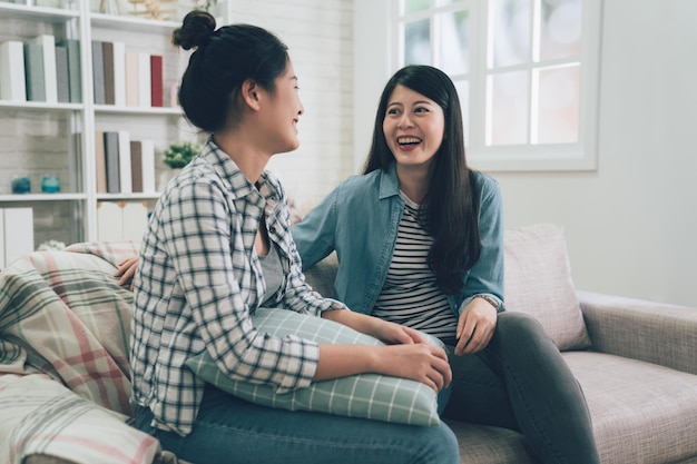 Close up photo of two asian young people excited tell speak news fresh gossips gladly smiling wear jeans sit on comfy sofa in living room. girls chatting talking on couch in bright room at home laugh