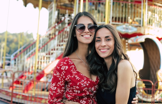 Close-up photo of two young good-looking female friends in festive dresses, posing in front of the carousel, looking at the camera and smiling with joy