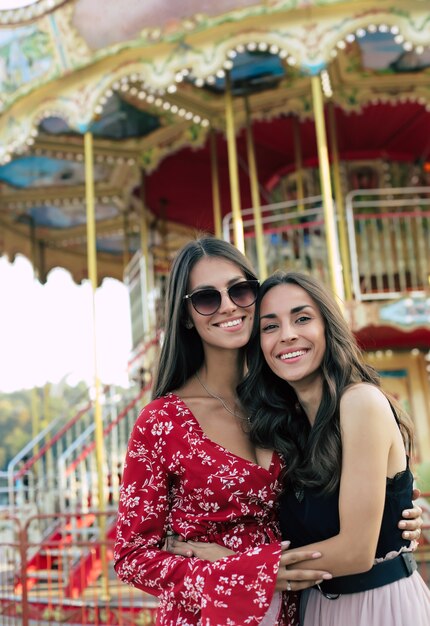 Close-up photo of two young good-looking female friends in festive dresses, posing in front of the carousel, looking at the camera and smiling with joy