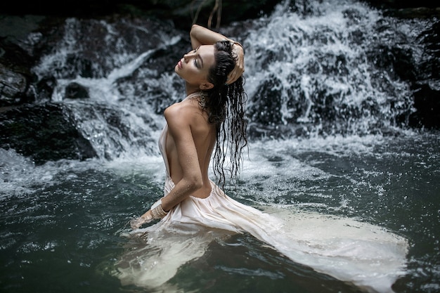 Close up photo of young beautiful woman in white dress is standing in the water on a big waterfall. 