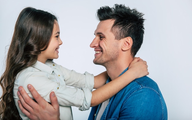 Close-up photo of a young dad and his little child, who are hugging tenderly and smiling, while looking in the camera together.