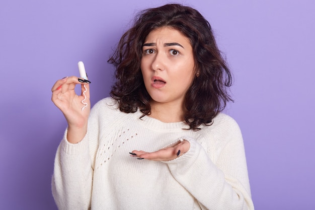 Close up photo of young dark haired female dresses white shirt holding tampon in hands