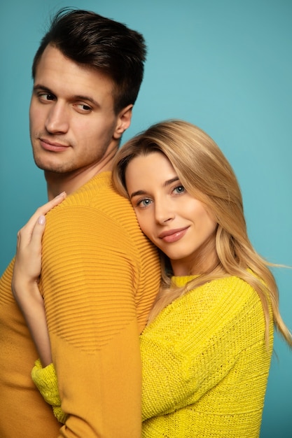 Close up photo of a young girl, who is standing in profile in a yellow outfit, hugging her boyfriend from the back, while he is trying to look at her.