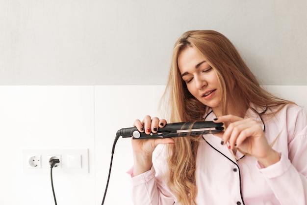 Close up photo of young lady sitting on bed and using hair straightener at home isolated