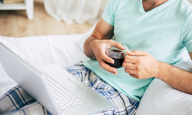 Close up photo of young man with a laptop and smartphone drinks coffee in bedroom.