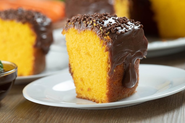 Close up piece of Brazilian carrot cake with chocolate frosting on wooden table with carrots in the background