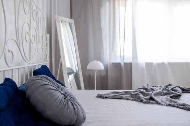 Close up of pillows and blanket on bed in bright bedroom interior with small lamp and mirror