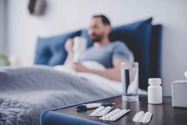 Крупный план таблеток на маленьком столике с больным мужчиной в постели, пьющим чай на заднем плане