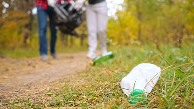 Foto primo piano di una bottiglia di plastica nell'erba sullo sfondo di una coppia che raccoglie immondizia nella foresta.