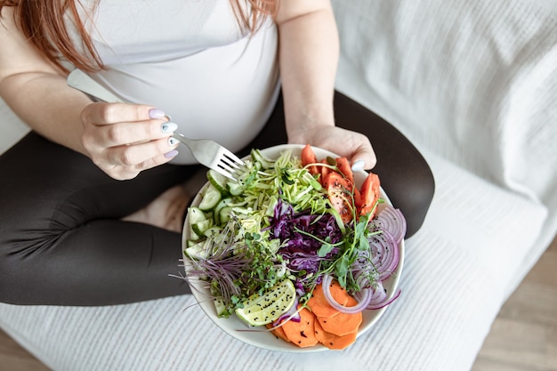 妊娠中の女性が手に取った新鮮な野菜の明るいサラダの付いたプレートのクローズアップ。