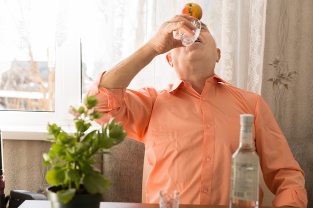 Close up Playful Elderly Drinking Vodka Wine with Apple on Forehead While Sitting at the Living Area Close to the Window.