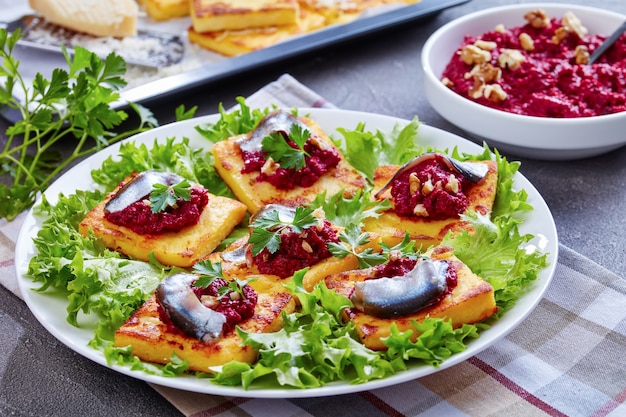 Close-up of Polenta Squares with Creamy beetroot puree, topped with anchovies and parsley on a white plate. freshly baked polenta bars and parmesan on a baking tray on a concrete table