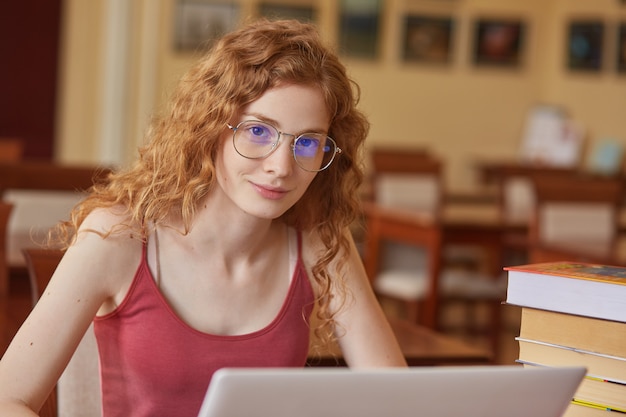 Close up portait of teenage woman sitting at table in front of opened lap top, wearing casual maroon t shirt