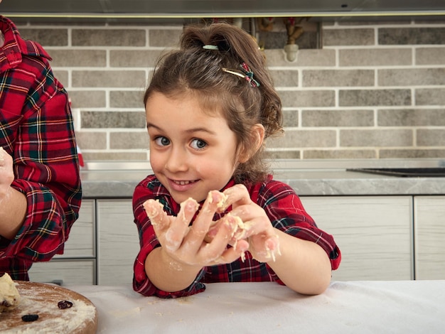 Close-up portrait of adorable gorgeous cute European little girl in red and green checkered dress playing with dough and smiling looking at camera