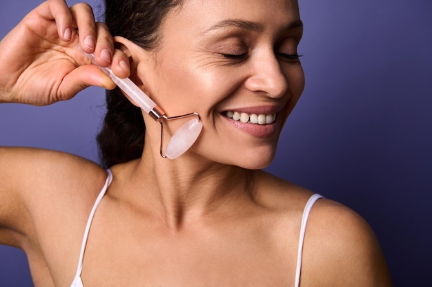 Close-up portrait of African American pretty woman doing face lifting smoothing anti-aging massage with pink jade stone roller, isolated over violet background with copy space. Face skin care concept