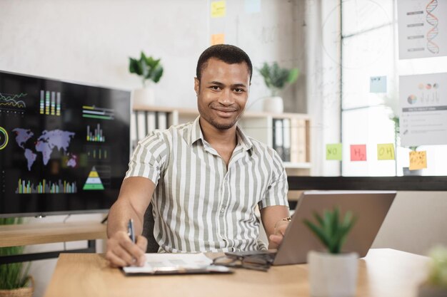 Photo close up portrait of african young male office manager in striped shirt writing financial report