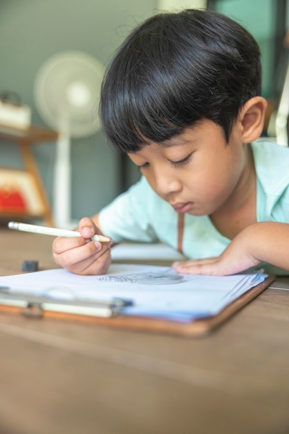 Close up portrait of Asian boy with black bangs black eyes with a smiling face wearing a light green and lying on the floor of his house drawing a picture of a girl with braids Education concept