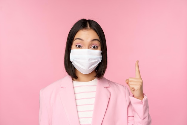 Close up portrait of asian businesswoman in medical face mask and suit pointing finger up showing advertisement top banner standing over pink background