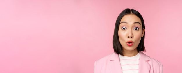 Close up portrait of asian corporate woman business lady looking surprised and amazed at camera standing in suit against pink background