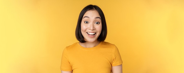 Close up portrait of asian girl showing surprised reaction raising eyebrows amazed reacting to big news standing over yellow background