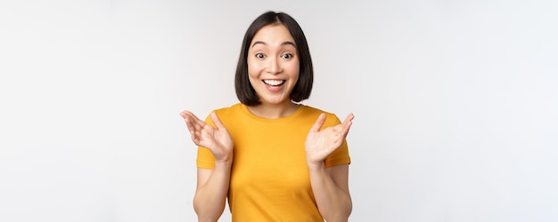 Close up portrait of asian woman looking surprised wow face staring impressed at camera standing over white background in yellow tshirt