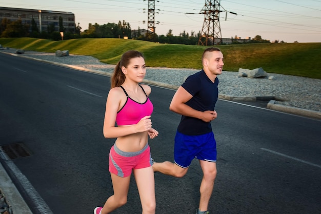 Close-up portrait of athletic couple running on the road, muscular build young runners working out while jogging in the park. Evening