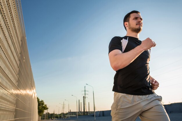 Close-up portrait of athletic man running along beautiful grey wall outdoors, muscular build young runner working out while jogging in the park. Sunset