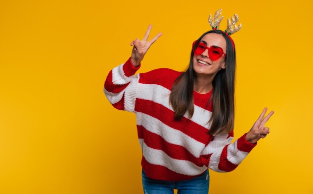 Close up portrait of an attractive excited smiling young girl in Christmas reindeer antlers and sweater while she posing isolated on yellow background in studio