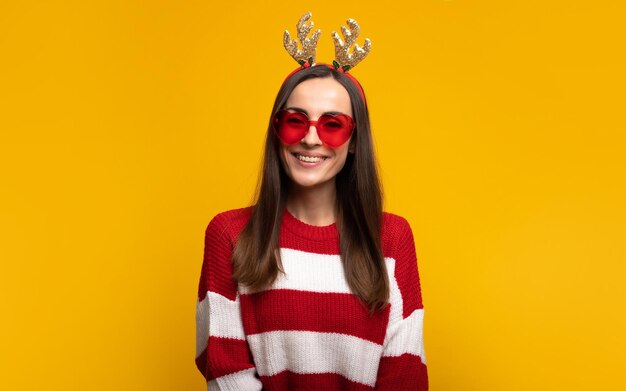 Close up portrait of an attractive excited smiling young girl in christmas reindeer antlers and sweater while she posing isolated on yellow background in studio