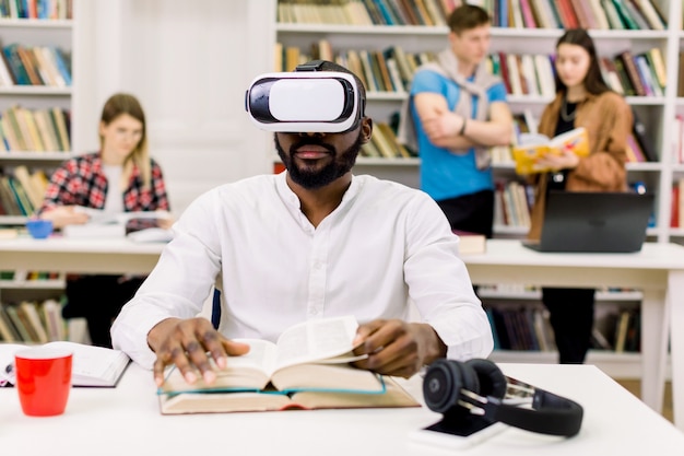 Close up portrait of attractive focused young african american bearded student sitting in library and reading book in vr goggles, while his groupmates talking on the space