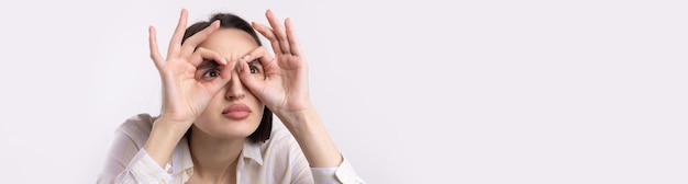 Close up portrait of attractive quirky young woman making binoculars with hands showing ok gesture on white studio background