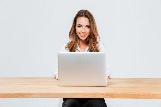 Close up portrait of an attractive smiling businesswoman using laptop while sitting at the office desk over white background