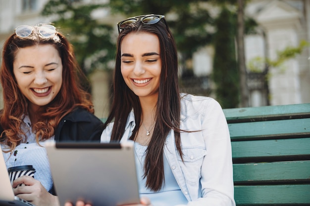 Close up portrait of a attractive woman with long dark hair looking to a tablet laughing sitting on a bench while her female friend is laughing