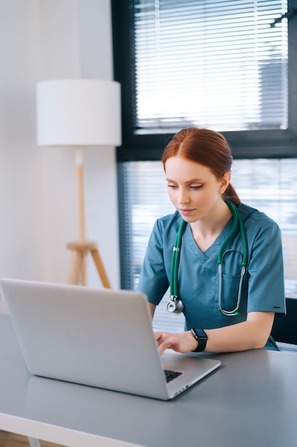 Close-up portrait of attractive young female practitioner doctor wearing blue green medical uniform working typing on laptop sitting at desk