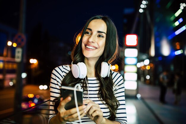 Close up portrait of attractive young happy woman in headphones with smart phone in hands outdoors