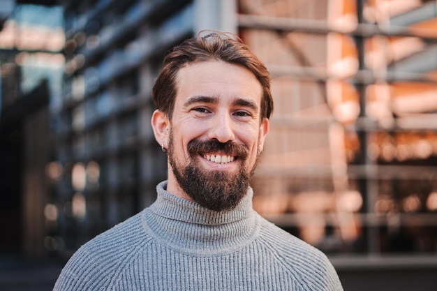 Close up portrait of awesome young caucasian male smiling and looking at camera at the background city office buildings