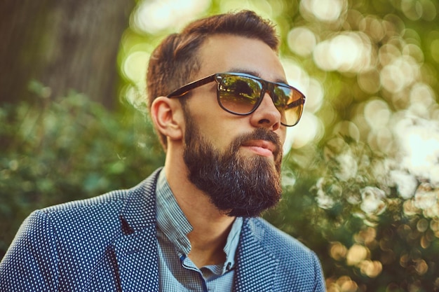 Close-up portrait of a bearded male with a stylish haircut, in a jacket and sunglasses, sitting on a bench in a city park.