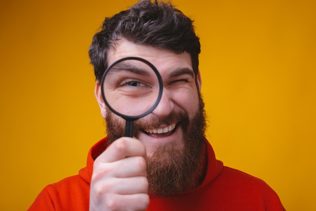 Close up portrait of a bearded man looking through a magnifying glass on yellow space.