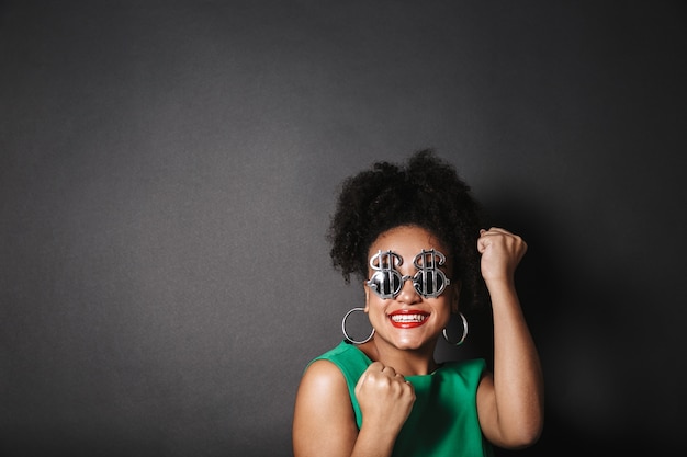 Close up portrait of a beautiful afro american woman wearing dollar shape sunglasses standing isolated over black space