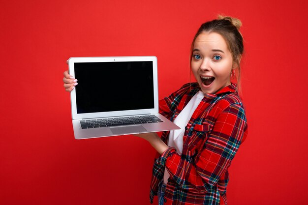 Close-up portrait of Beautiful amazed young woman holding laptop wearing red shirt looking at camera isolated on red background. Empty space, cutout