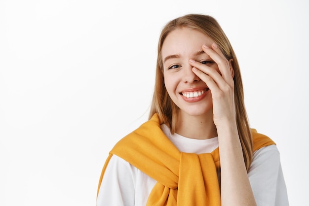 Close up portrait of beautiful blond girl touching fresh glowing healthy skin smiling white teeth and looking happy at camera standing against studio background