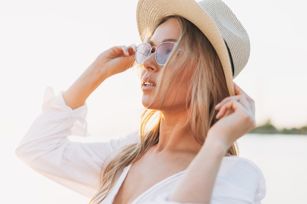 Close up portrait of beautiful blonde young woman in white shirt and straw hat on the sunset